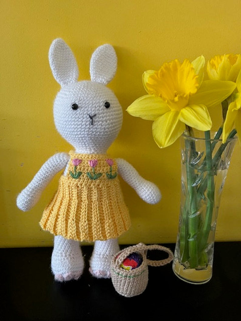 Pode incluir: Um coelho de croch&ecirc; branco vestindo um vestido amarelo com flores bordadas em rosa e verde. O coelho est&aacute; segurando uma pequena cesta de croch&ecirc; marrom com ovos de P&aacute;scoa coloridos dentro. O coelho est&aacute; em p&eacute; na frente de um vaso com narcisos amarelos.