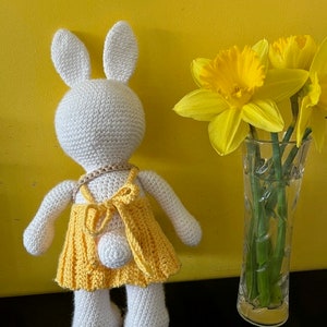 Pode incluir: Um coelho de croch&ecirc; branco vestindo um vestido de croch&ecirc; amarelo. O coelho est&aacute; em p&eacute; sobre uma superf&iacute;cie preta em frente a uma parede amarela. H&aacute; narcisos amarelos em um vaso de vidro atr&aacute;s do coelho.