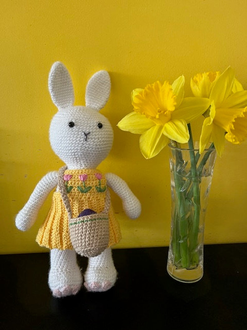 White crocheted rabbit wearing a lemon yellow dress decorated with pink flowers. She has a basket with a strap which goes across her body and is big enough to hold a chocolate egg.