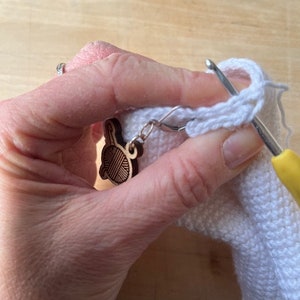 Pode incluir: Um close-up de uma m&atilde;o segurando uma pe&ccedil;a de croch&ecirc; branca com um gancho de croch&ecirc;. Um amuleto de madeira em forma de novelo de l&atilde; est&aacute; preso &agrave; pe&ccedil;a de croch&ecirc;.