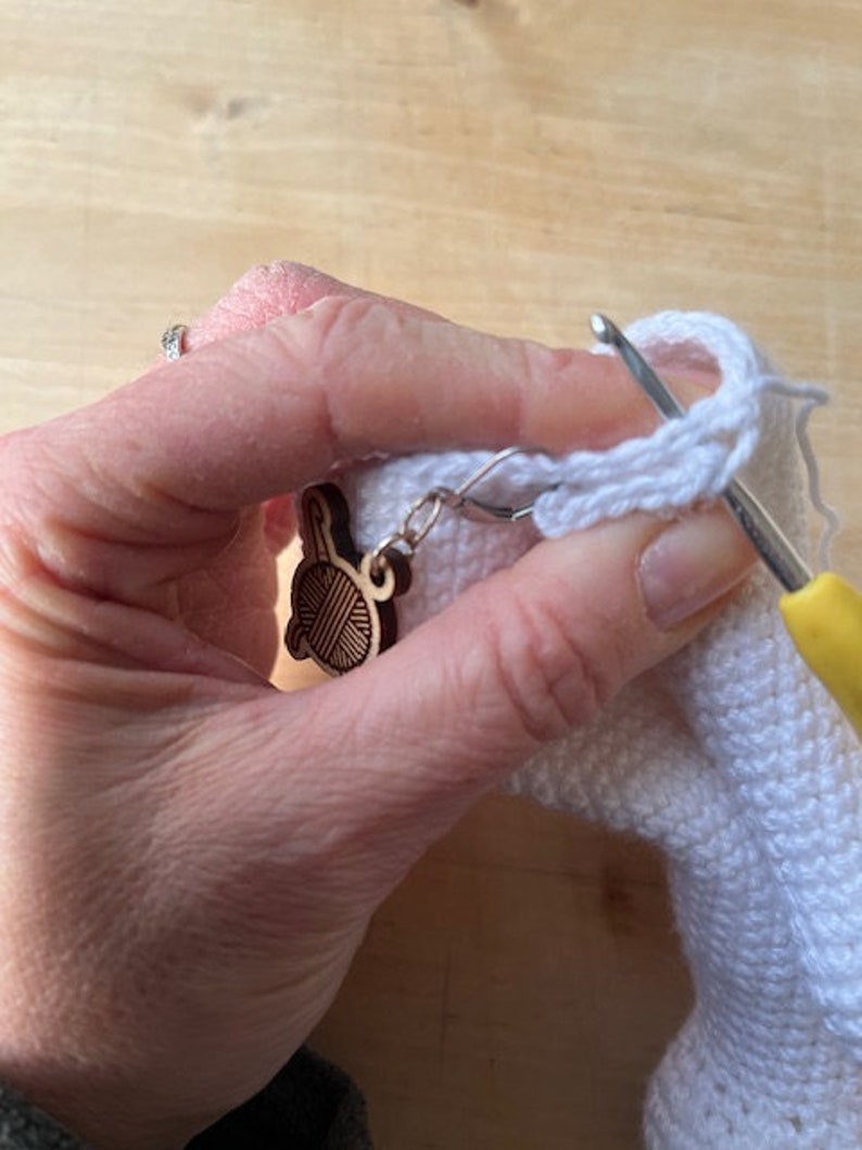 Pode incluir: Um close-up de uma m&atilde;o segurando uma pe&ccedil;a de croch&ecirc; branca com um gancho de croch&ecirc;. Um amuleto de madeira em forma de novelo de l&atilde; est&aacute; preso &agrave; pe&ccedil;a de croch&ecirc;.