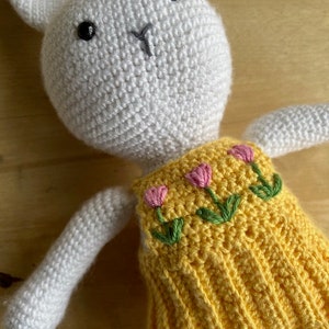 Pode incluir: Um coelho de croch&ecirc; branco vestindo um vestido amarelo com flores bordadas de rosa. O coelho tem olhos pretos e um nariz costurado.