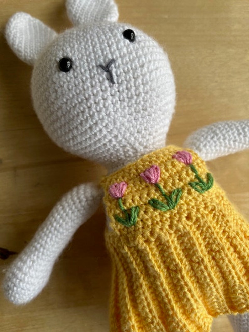 Pode incluir: Um coelho de croch&ecirc; branco vestindo um vestido amarelo com flores bordadas de rosa. O coelho tem olhos pretos e um nariz costurado.