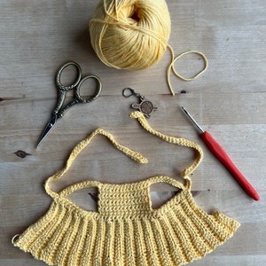 Pode incluir: Um top de croch&ecirc; amarelo com duas al&ccedil;as, uma bola de fio amarelo, um par de tesouras, um gancho de croch&ecirc; e um pequeno amuleto de metal.
