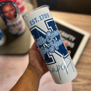 以下が含まれることがあります： 「EST. 1789」と「North Carolina Tar Heels」の文字が入った白と青のタンブラー。タンブラーには銀色の蓋と、底に青色のサインが入っています。円筒形のタンブラーはスポーツをテーマにしています。