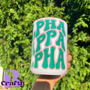 May include: White tumbler with the text "ALPHA" in green, outlined in pink. The tumbler is held in front of a green leafy background. The bottom left corner has a purple logo with the text "Crafty Kreations".