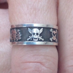 Può includere: Un anello argentato con una fascia scura con disegni a tema pirata. L'anello presenta motivi di teschio e ossa incrociate, insieme ad altri simboli pirata. L'anello ha una finitura lucida.