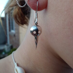 May include: Silver skull-shaped earring with a pointed bottom. The earring is hanging from a silver hook.