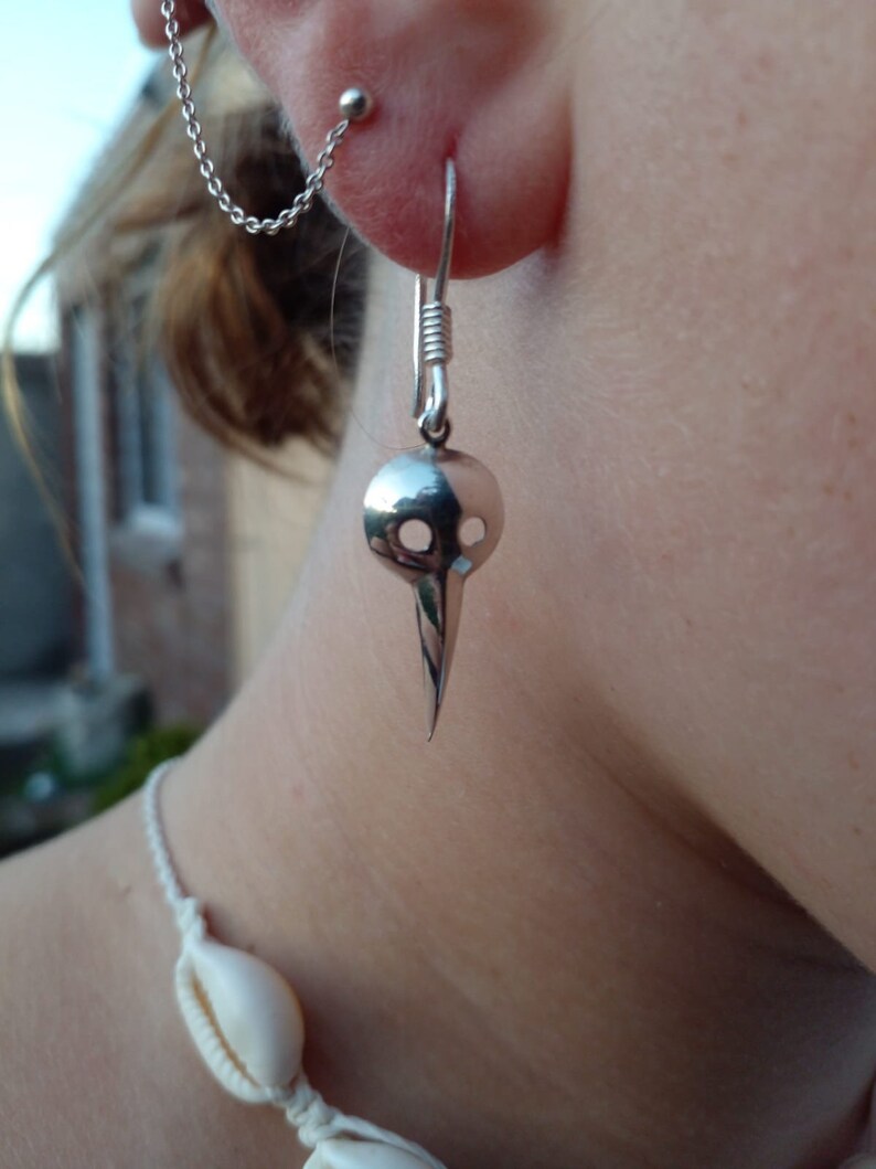 May include: Silver skull-shaped earring with a pointed bottom. The earring is hanging from a silver hook.