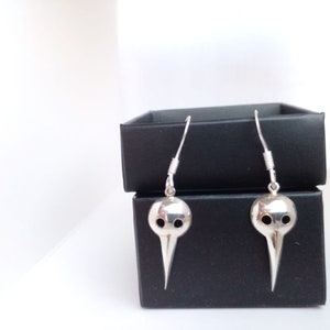 May include: A pair of silver earrings shaped like a bird's head with two holes for eyes. The earrings are hanging from a black box.