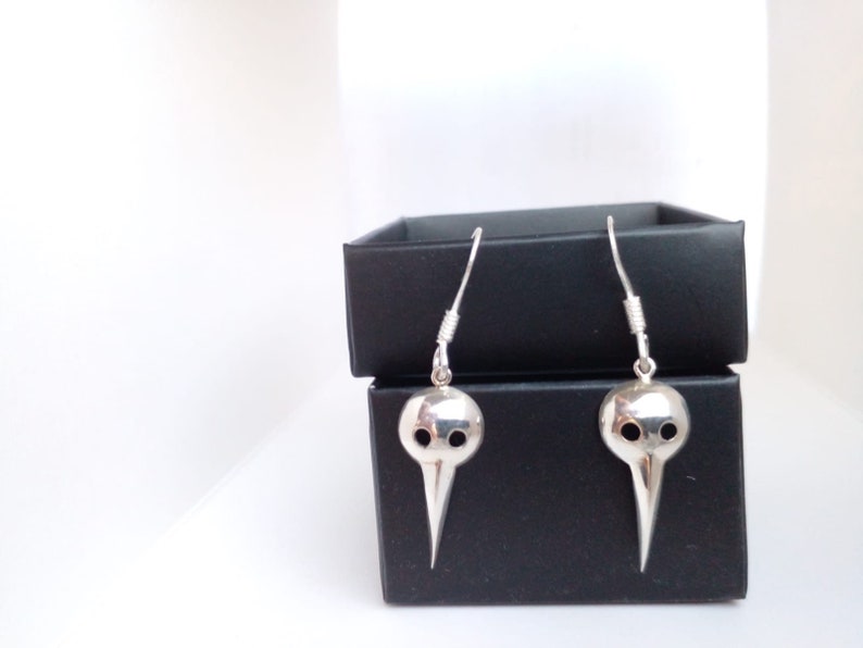 May include: A pair of silver earrings shaped like a bird's head with two holes for eyes. The earrings are hanging from a black box.
