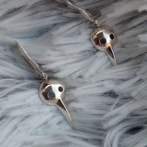 May include: A pair of silver earrings with a bird mask design. The masks have a long, pointed beak and two small holes for eyes.