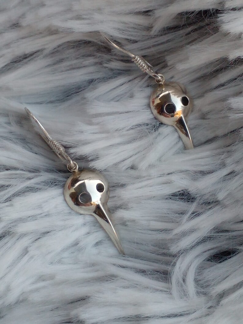 May include: A pair of silver earrings with a bird mask design. The masks have a long, pointed beak and two small holes for eyes.