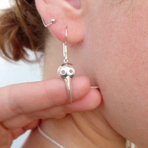 May include: Silver bird skull dangle earrings with a long, pointed beak. The earrings are hanging from a person's ear.
