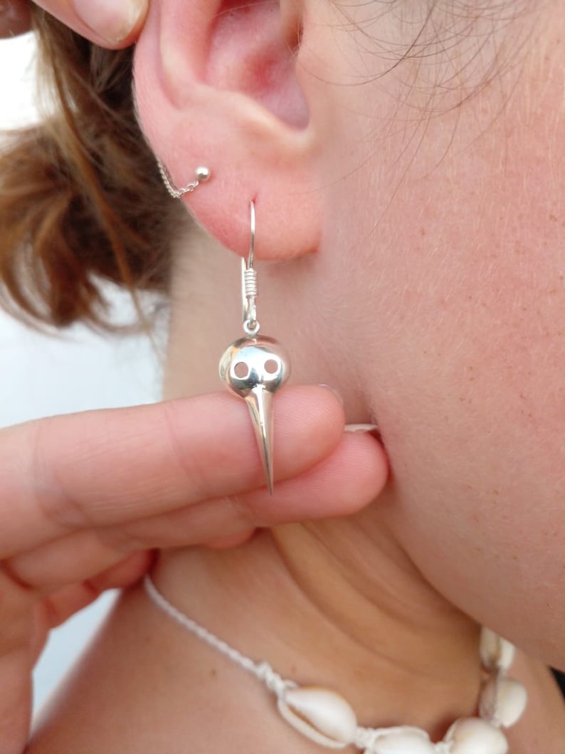 May include: Silver bird skull dangle earrings with a long, pointed beak. The earrings are hanging from a person's ear.
