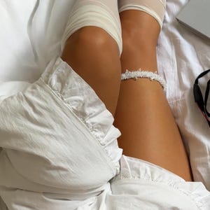 May include: A white beaded garter featuring small clear crystals and pearls arranged in a delicate, pointed pattern. The accessory is worn around the thigh, paired with a white ruffled garment and sheer white hosiery, creating a layered look.