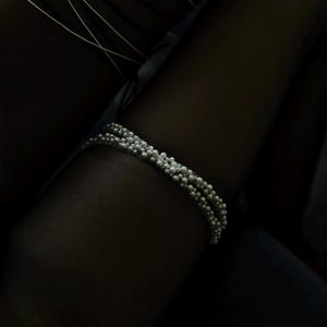 May include: A silver bracelet with three strands of small, round beads. The bracelet is set against a dark background, highlighting its delicate design. The beads have a textured appearance, adding visual interest to the jewelry.