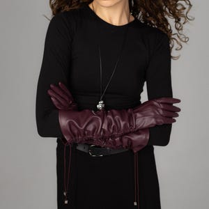 Puede incluir: Guantes largos de cuero burdeos con diseño fruncido. Los guantes se extienden más allá de los codos y tienen cordones en la muñeca. La modelo lleva una camiseta negra de manga larga y un cinturón negro.