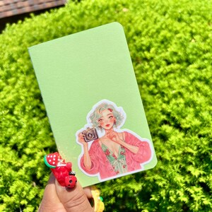 May include: A light green notebook with a sticker of a woman holding a camera. The woman has light blue hair and is wearing a pink jacket and floral dress. The notebook is held in front of a green bush.