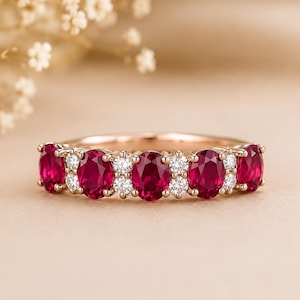 Könnte beinhalten: Ein Ring aus Roségold mit vier oval geschliffenen, tiefroten Rubinen, die sich mit Paaren kleiner, rund geschliffener weißer Diamanten abwechseln. Die Ringschiene besteht aus poliertem Roségold und zeigt eine symmetrische Anordnung der Edelsteine.