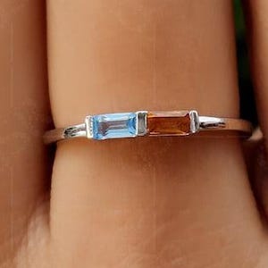 Könnte beinhalten: Ein silberner Ring mit zwei rechteckigen Edelsteinen: einem hellblauen und einem bernsteinfarbenen. Die Edelsteine sind nebeneinander auf einem schmalen Band angeordnet. Der Ring wird an einem Finger gezeigt.