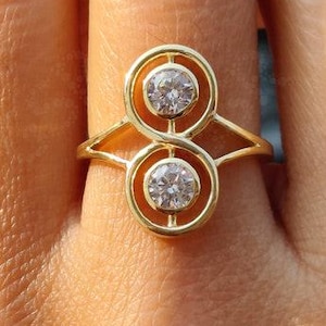 May include: A gold ring with two clear, round gemstones set in circular frames. The ring has a connecting band and a gold band. The ring is displayed on a person's finger.