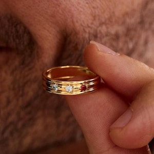 Pode incluir: Um anel de banda dourada com três pequenas pedras cravadas, dispostas horizontalmente no centro. As pedras consistem numa gema azul-clara à esquerda, uma gema branca transparente no meio e uma gema vermelho-escura no lado direito da banda.