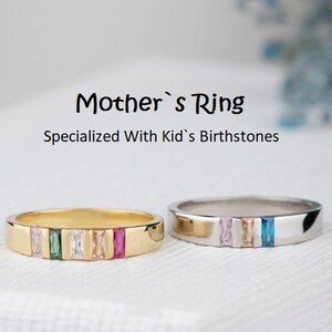 May include: Two rings with colourful gemstones. One ring is gold-toned, the other is silver-toned. The rings are inscribed with the text "Mother's Ring" and "Specialized With Kid's Birthstones". The rings have a band of rectangular gemstones in various colours.