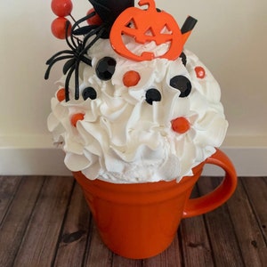 Puede incluir: Taza de cerámica naranja con crema batida blanca, caramelos negros y naranjas, una araña negra, un lazo negro y un recorte de calabaza naranja en la parte superior.