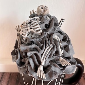 Pode incluir: Caneca de cerâmica preta com um design texturizado cinza com tema de Halloween. O design apresenta um esqueleto com um crânio no topo de uma pilha de tecido cinza. A caneca tem a palavra "Witch" escrita na frente.