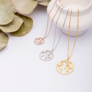 May include: Three gold necklaces with a world map pendant. The necklaces are on a white surface with green leaves in the background. The necklaces are different lengths and are made of gold, silver, and rose gold.