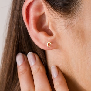 May include: A close-up of a gold-colored stud earring shaped like the letter "C" in a person's ear. The earring is small and simple, with a polished finish. The person's skin is fair, and their hair is brown.