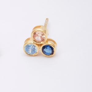 May include: Gold stud earrings with three round gemstones. The gemstones are light blue, pink, and dark blue.