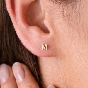 May include: A close-up of a person's ear with a small, gold-colored stud earring. The earring is in the shape of the letter "M". The skin tone is light, and the earring is centered on the earlobe.