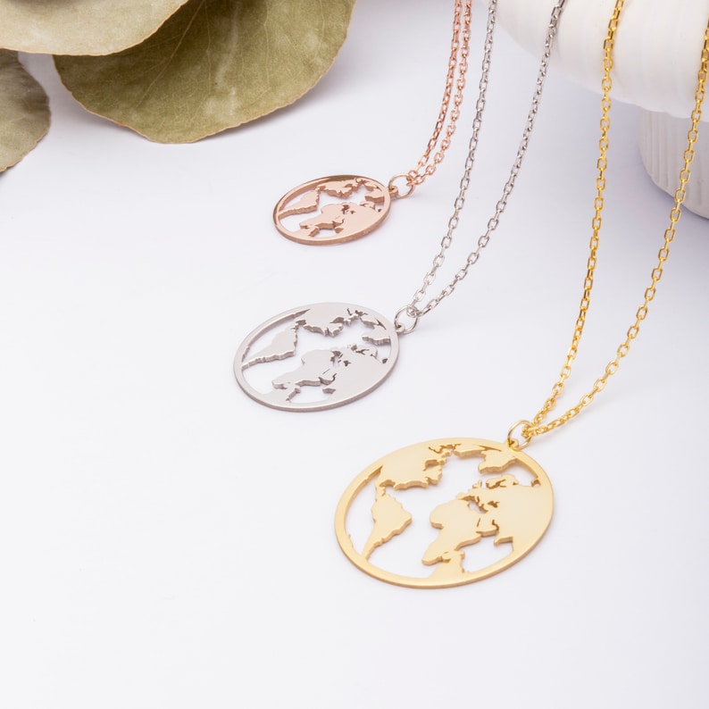 May include: Three gold necklaces with a world map pendant. The necklaces are on a white surface with green leaves in the background. The pendants are different sizes and are made of gold, silver, and rose gold.