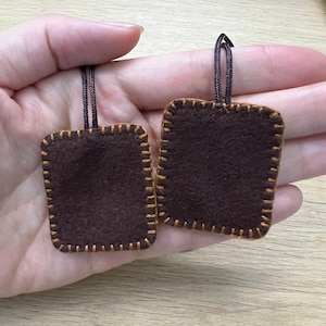 Puede incluir: Dos adornos cuadrados de fieltro marrón con costuras doradas en los bordes. Cada adorno tiene un lazo de cordón marrón oscuro en la parte superior. Los adornos se muestran sobre un fondo claro.