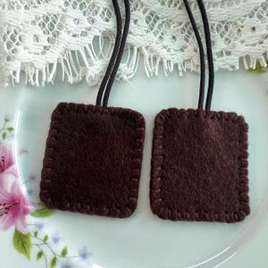 Pode incluir: Dois remendos quadrados de feltro castanho-escuro com cantos arredondados, cada um com uma borda de ponto caseado espessa na cor correspondente. Cada remendo está preso a um laço de cordão fino castanho-escuro que sai da borda superior.