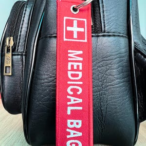 May include: A red fabric tag with the words "MEDICAL BAG" in white, and a white cross symbol, attached to a silver keyring. The tag is hanging from a black leather backpack with silver zips.