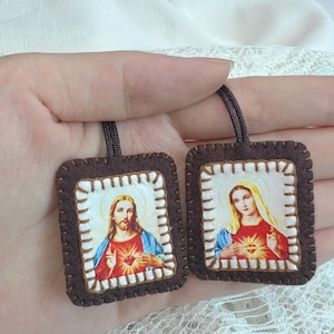 May include: Two brown felt religious pendants with images of Jesus and Mary. Each pendant has a stitched border and a cord for hanging. The images feature Jesus with a red heart and Mary with a red heart.