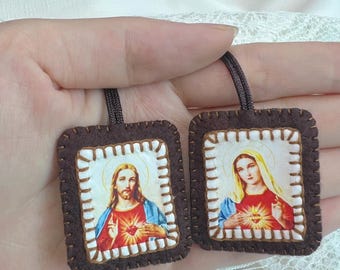 Puede incluir: Dos colgantes cuadrados de tela con imágenes religiosas. Uno representa el Sagrado Corazón de Jesús y el otro el Inmaculado Corazón de María. Cada uno tiene un borde de fieltro marrón oscuro con puntadas blancas y un cordón marrón arriba.