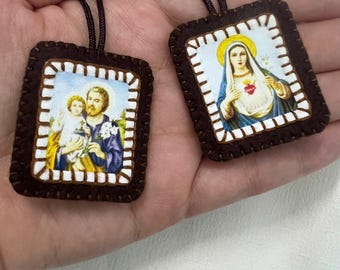 Può includere: Due ciondoli rettangolari con immagini religiose. Uno mostra una figura che tiene in braccio un bambino con fiori bianchi, l'altro una figura con un'aureola gialla e un cuore rosso. Entrambi hanno bordi in feltro marrone scuro con cuciture bianche e cordini neri.