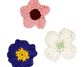 May include: Three crocheted flowers in pink, white, and blue. The pink flower has a maroon centre, the white flower has a green centre, and the blue flower has a yellow centre.