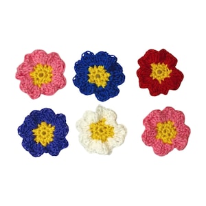 Op de afbeelding: Zes gehaakte bloemen in verschillende kleuren, waaronder roze, blauw, rood en wit. Elke bloem heeft een geel centrum.