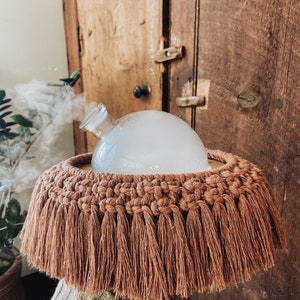 Op de afbeelding: Een glazen aromatherapie-diffuser met een bolvormige bovenkant, die damp afgeeft, staat op een macrame franjebasis. De basis heeft een warme, aardse bruine kleur. De achtergrond toont een houten deur.