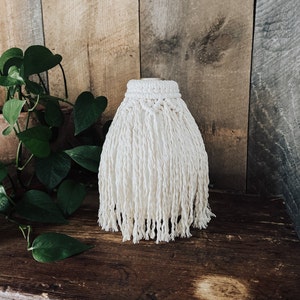May include: A white macrame vase with a fringe design. The vase is made of natural fibers and has a rustic, bohemian style.