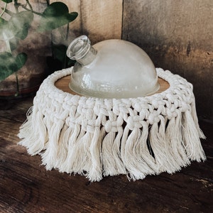 May include: A clear glass diffuser with a white macrame base and fringe. The diffuser is emitting steam.