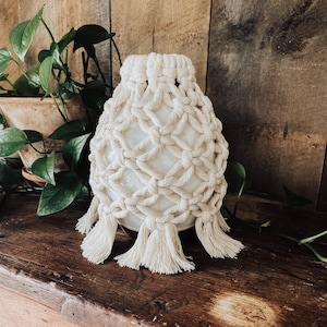 Puede incluir: Un difusor de cerámica blanco con una cubierta de macramé de crochet. La cubierta tiene un fleco alrededor de la parte inferior y está hecha de algodón natural de color crema.