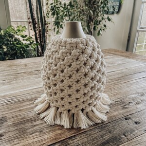 May include: A white crocheted lampshade with a fringe trim. The lampshade is sitting on a wooden table.