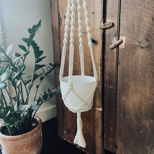 May include: A white macrame plant hanger with a white ceramic pot hanging from an antique wooden cabinet.