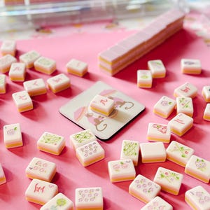 May include: A collection of Mahjong tiles with a pink and yellow color scheme, scattered on a pink surface. The tiles feature various symbols and floral designs. A matching tile rack and a coaster with a monkey design are also present.